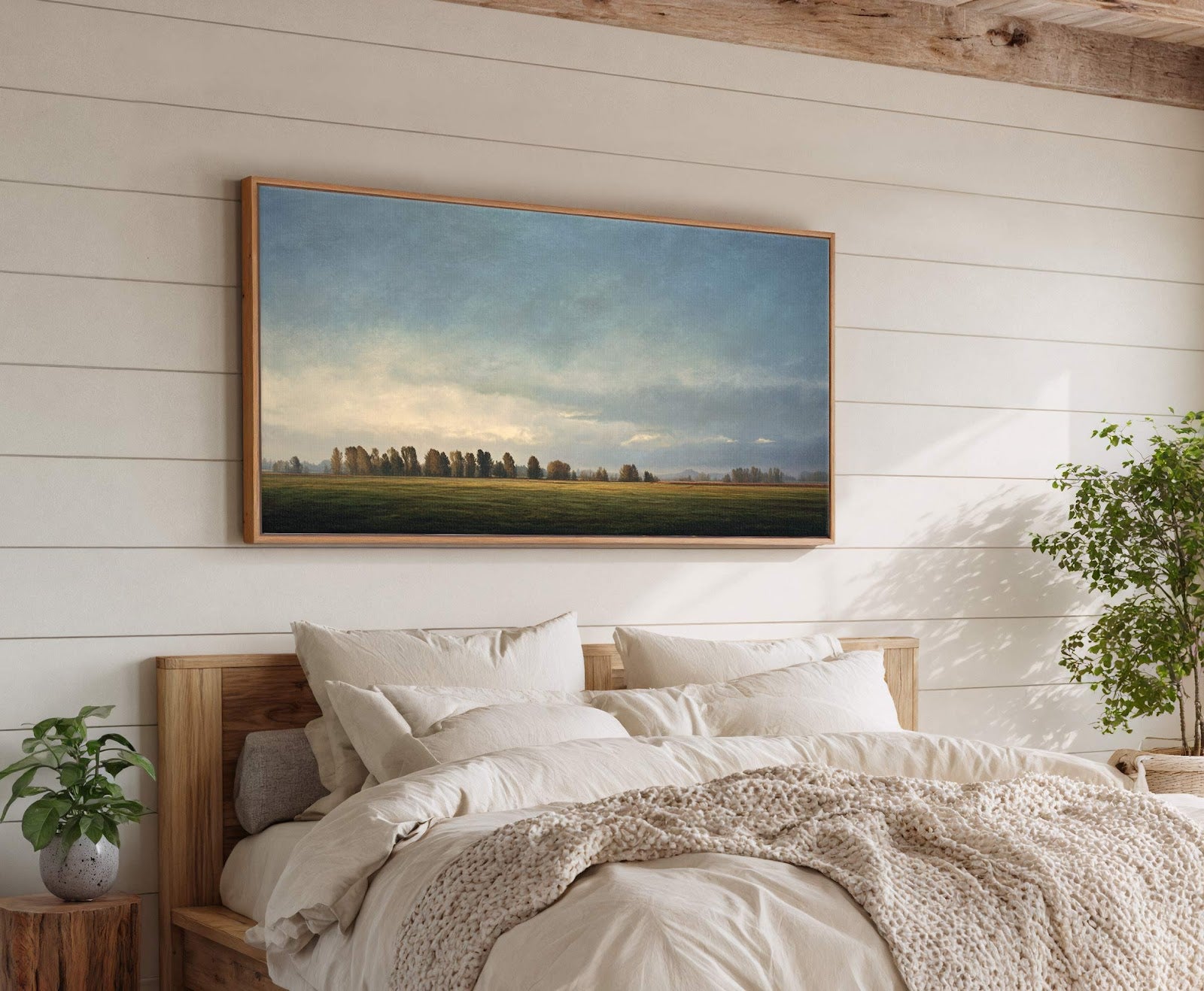 A horizontal canvas print artfully depicts a serene landscape with a treeline and fields under a moody, cloudy sky, showcasing a muted palette and painterly style, displayed above a bed.