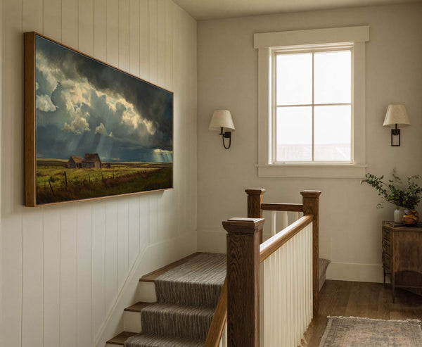 This horizontal canvas wall art depicts a dramatic landscape with stormy clouds and sunbeams illuminating rustic barns, set against a neutral wall above a staircase and next to a window.
