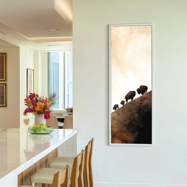 This vertical canvas wall art features a watercolor-style depiction of bison silhouettes ascending a textured, earthy brown hill against a soft beige background, framed in white.