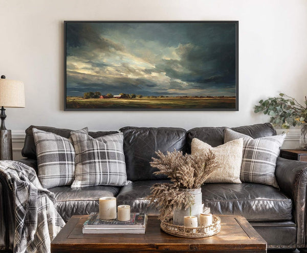 This landscape painting print features a dramatic, cloudy sky over a rural scene with barns. It is a horizontal canvas in a black floater frame hanging above a dark leather.