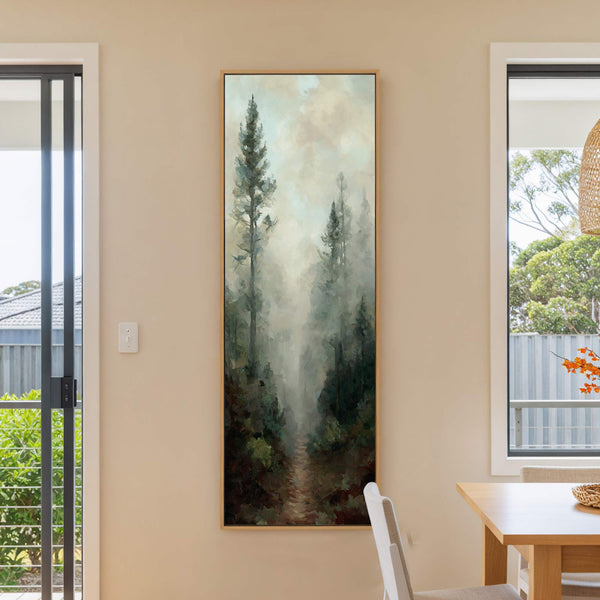 This vertical, impressionistic painting print showcases a foggy forest path with a muted green and brown palette, framed in natural wood, hanging on a beige wall next to a window.