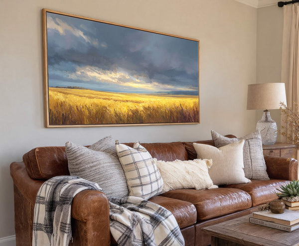 This horizontal canvas wall art depicts a dramatic, impressionistic landscape of a golden wheat field under a stormy, cloudy sky, framed in natural wood, hanging above a brown leather sofa.