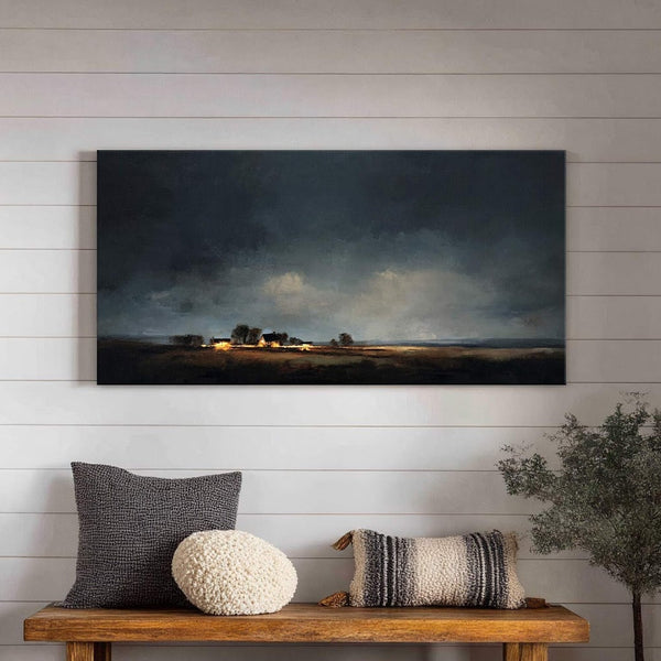 This horizontal canvas wall art depicts a dimly lit landscape with a cluster of illuminated homes against a stormy sky, rendered in a moody, impressionistic style, displayed above a wooden.