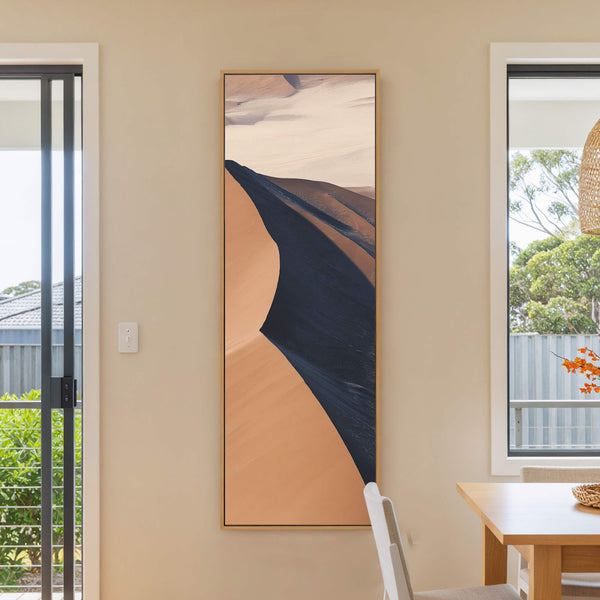 This vertical canvas wall art features a desert landscape in a warm, earthy palette with a minimalist, abstract style. It is displayed on a neutral wall in a room with.
