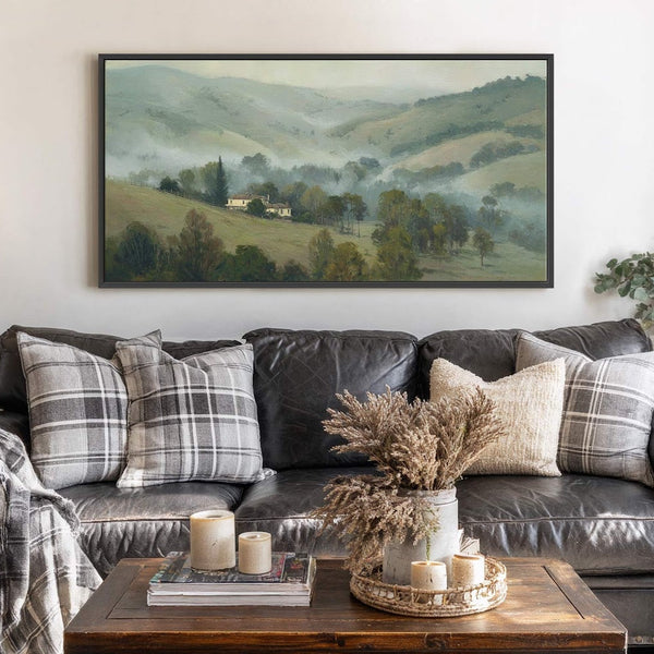 This horizontal, framed canvas wall art depicts a misty, rolling landscape with a house and trees in muted green and gray tones, displayed above a dark leather sofa with plaid.