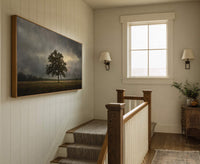 A horizontal canvas print with a moody palette depicts a solitary tree in a field under a dramatic, cloudy sky, framed in natural wood, hanging on a wall with vertical.