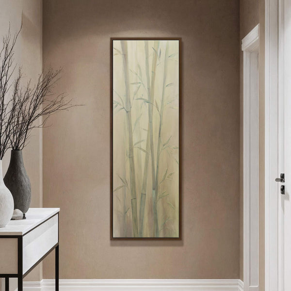 A vertical, painterly art print of pale green bamboo stalks against a soft, pale yellow background is framed in a warm brown floater frame, displayed on a taupe wall above.