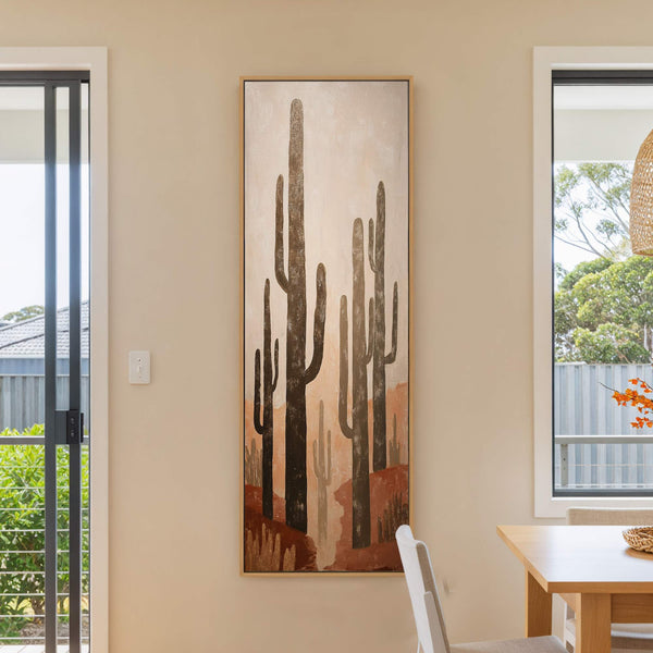 This vertical, stretched canvas wall art features a desert landscape with saguaro cacti in a rustic, earthy palette, displayed on a light beige wall next to a window and a.