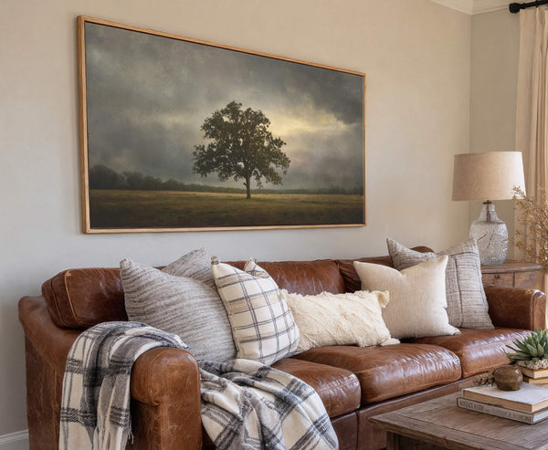 This horizontal canvas wall art features a lone oak tree in a field under a dramatic, moody sky. The artwork, with its muted, earthy palette, is framed in natural wood.