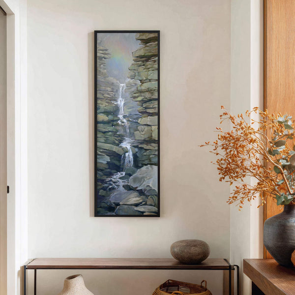 This vertical canvas wall art features a serene waterfall cascading over rocky cliffs, rendered in a muted palette with hints of rainbow mist, framed in black and displayed on a.