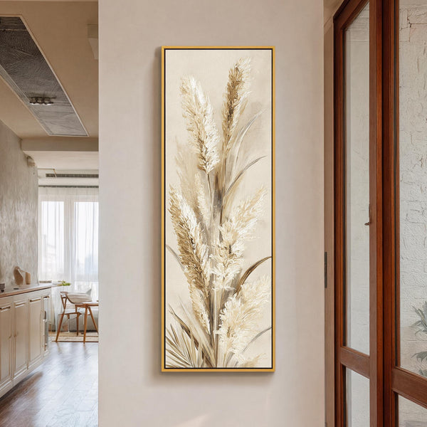 This vertical canvas print features textured pampas grass in a painterly style and neutral palette, displayed in a gold floater frame on an off-white hallway wall near a window.