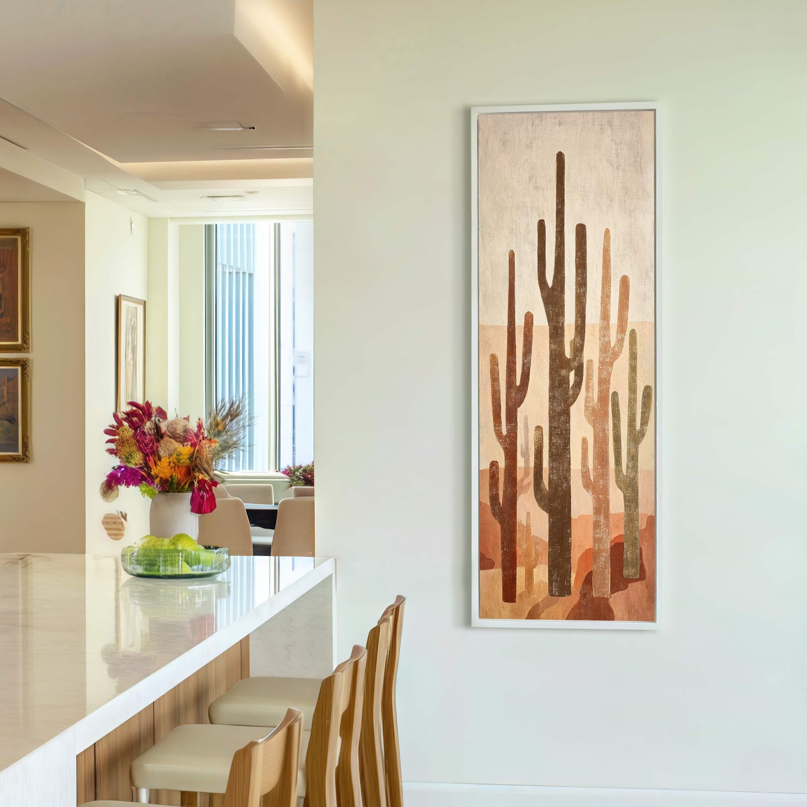A vertical canvas wall art print featuring a stylized desert landscape with saguaro cacti in earthy tones, framed in white, hangs on a plain wall in a room with a.