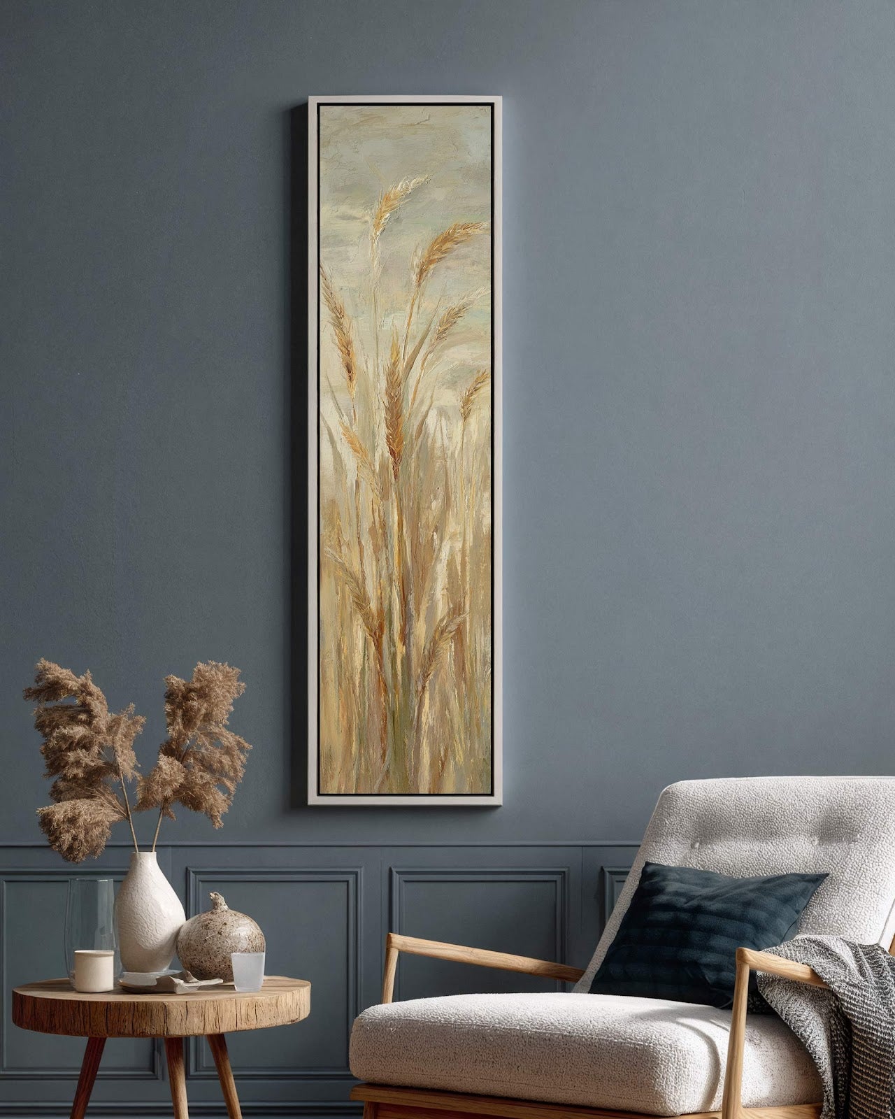 This vertical canvas print depicts textured wheat in a beige and gold palette, set in a white floater frame on a blue-gray paneled living room wall near an armchair.
