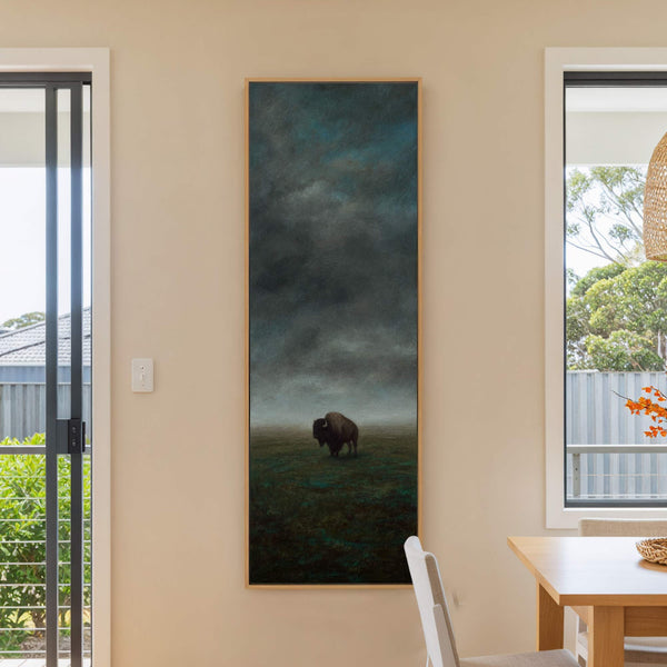 A vertical canvas print depicts a lone bison in a moody landscape with a dark, cloudy sky and a muted green field, framed in natural wood, displayed on a beige.