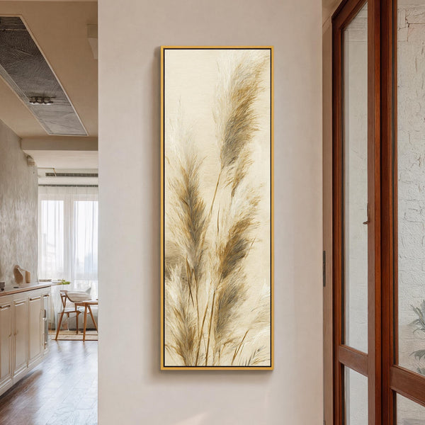 This vertical canvas wall art depicts textured pampas grass in beige tones, showcased in a gold floater frame on a cream hallway wall beside a wooden door.