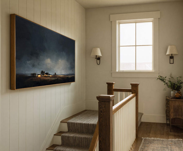 This horizontal canvas wall art displays a dark, moody landscape with illuminated houses in a painterly style. It is set against a creamy, vertical shiplap wall in a hallway, near.