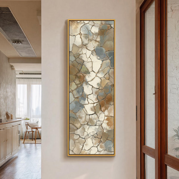 This vertical abstract canvas print features a textured mosaic pattern in earthy tones, showcased in a gold floater frame on a cream hallway wall beside a glass-paneled door.