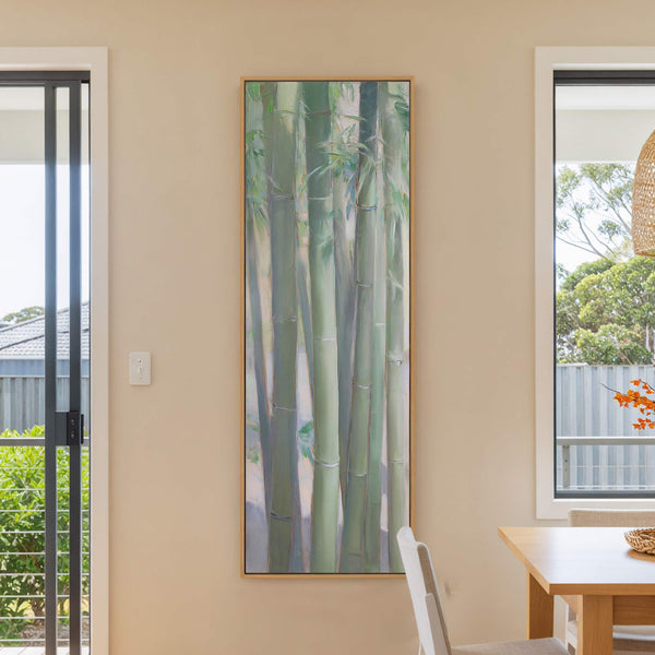 This vertical, impressionistic canvas wall art features a soothing palette of greens and blues depicting bamboo stalks. It is displayed on a cream-colored wall in a room with a wooden.