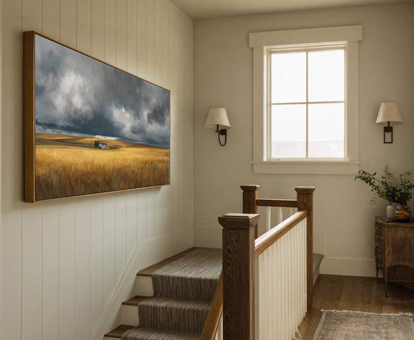 This horizontal canvas wall art features a dramatic stormy sky over a golden field with a small house and trees, painted in a textured, impressionistic style and framed in natural.