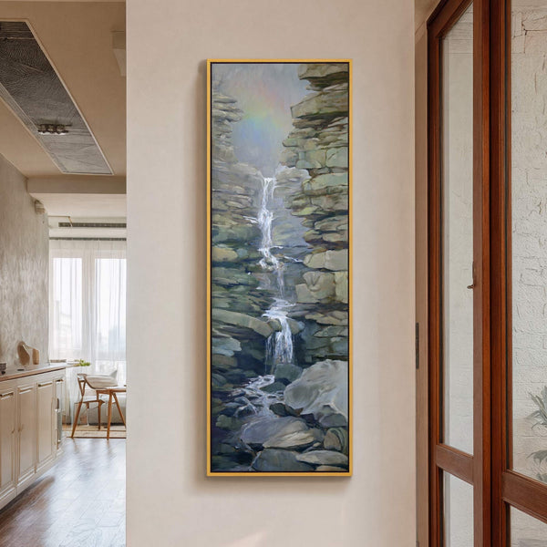 This vertical, rectangular canvas wall art features a realistic painting of a waterfall cascading through rocky terrain, using a cool, muted palette and framed in gold. It hangs on a.