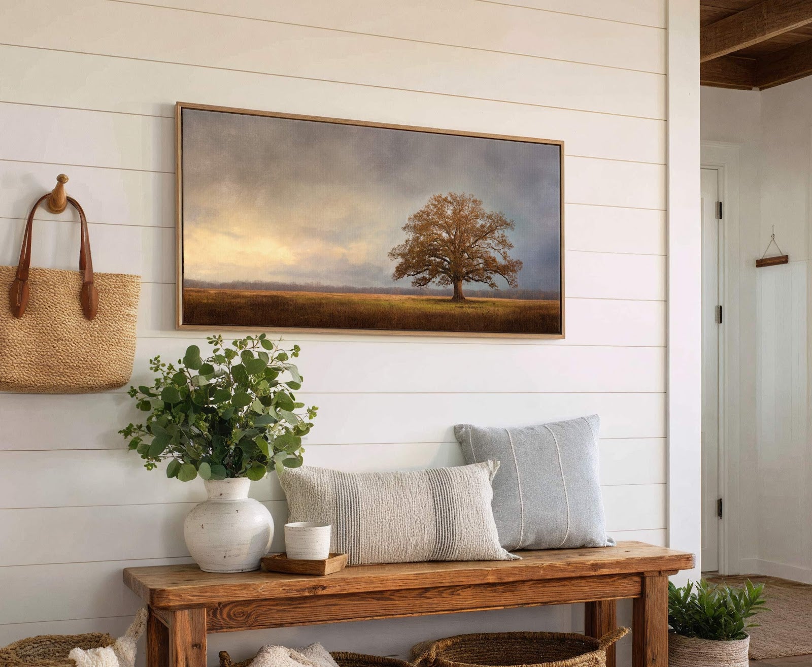 This horizontal canvas wall art features a textured painting of a solitary tree in a field under a dramatic sky, displayed above a wooden bench with pillows and a vase.