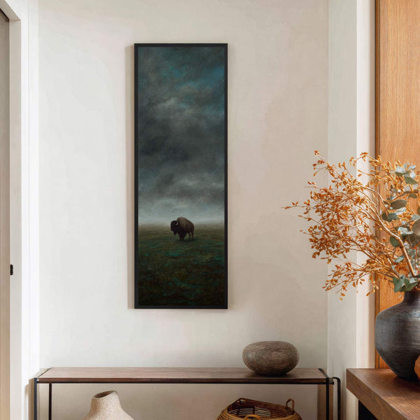 This vertical, framed canvas print features a lone bison in a misty, moody landscape with a dark, moody palette. It hangs on a white wall above a narrow console table.