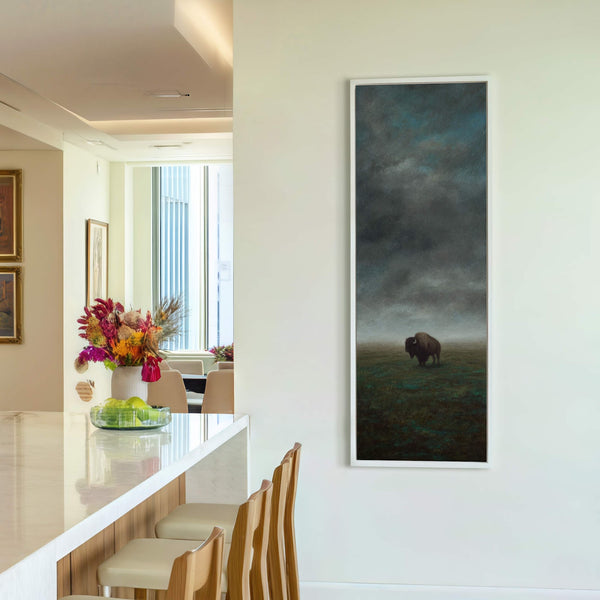 A vertical canvas print of a solitary bison in a misty field features a dark, moody palette and a painterly style, displayed in a white floater frame on a neutral.