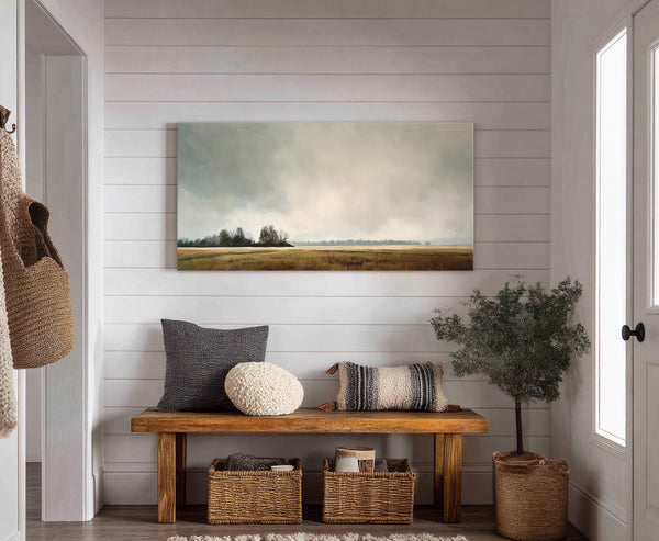 A horizontal canvas print displays a pastoral landscape with a muted palette and soft brushstrokes, set against a white paneled wall above a wooden bench with cushions and baskets.