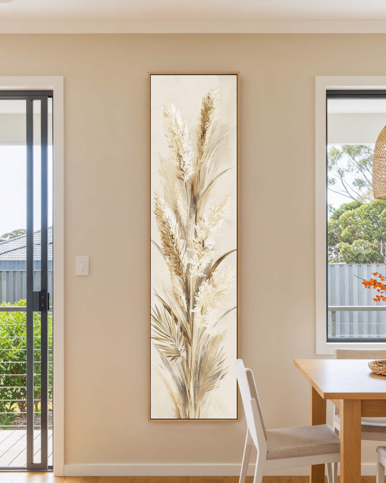 This vertical canvas print showcases painterly pampas grass in neutral beige tones, mounted in an oak floater frame on a tan dining room wall beside a light wooden table.
