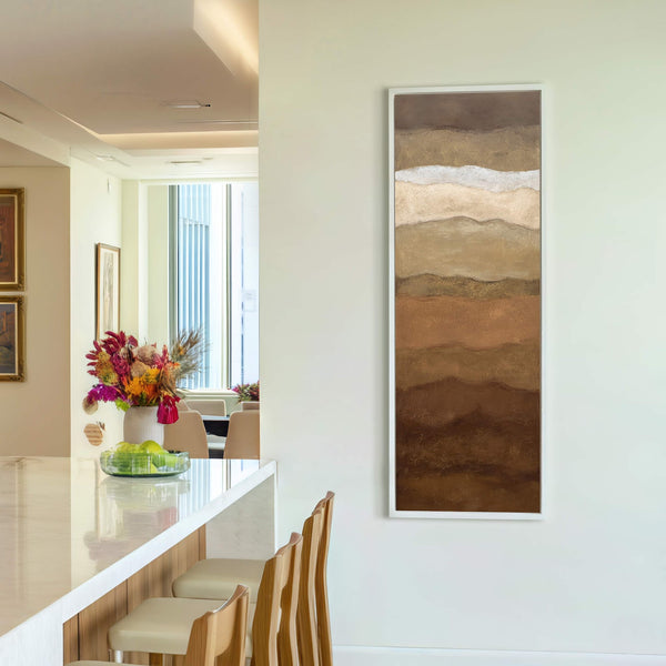 This vertical canvas wall art features abstract layered earth tones in a textured style, framed in white and displayed on a light cream wall above bar stools.