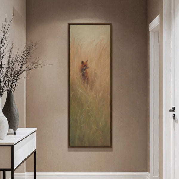 A vertical canvas print displays a fox in tall grass with a warm, impressionistic palette and a walnut floater frame, hanging on a tan wall near a console with a.