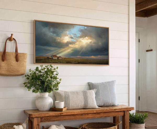 This horizontal canvas wall art depicts a dramatic landscape of houses under a cloudy sky with sunbeams. It is framed in natural wood and hangs on a white shiplap wall.