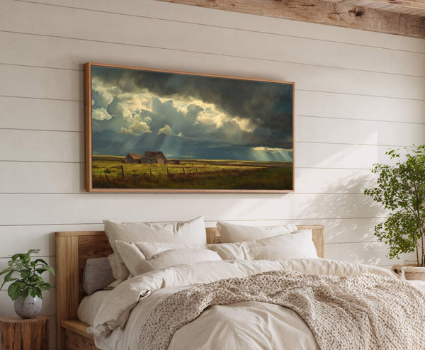 This horizontal canvas wall art features a dramatic pastoral landscape painting with a dark, stormy sky and sunbeams illuminating old barns. It is displayed above a bed in a room.