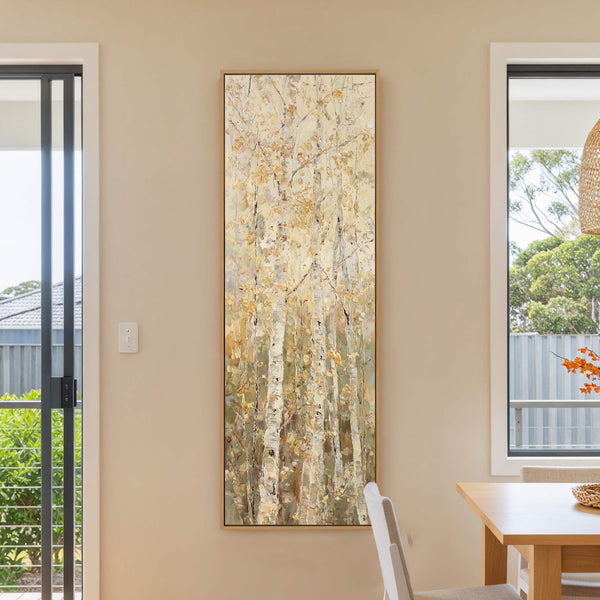 This vertical canvas wall art features a textured, impressionistic depiction of birch trees with a warm, golden palette, set against a neutral beige wall between two windows with a table.