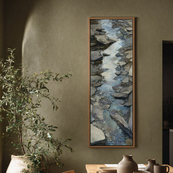 This vertical impressionistic painting print of a rocky stream features earthy tones, displayed in an oak floater frame on an olive textured living room wall beside a large plant.