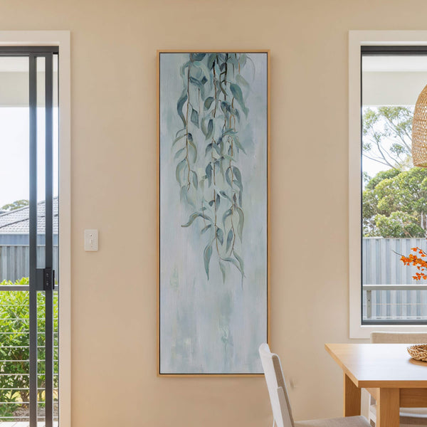 This vertical canvas wall art features a muted palette of blues and greens with a painterly style depicting weeping eucalyptus leaves. It is displayed on a beige wall in a.