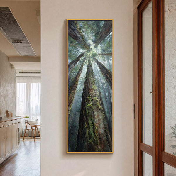 This vertical canvas wall art depicts a forest canopy from below with a palette of greens and browns, framed in gold and hung on a cream wall near a wooden.