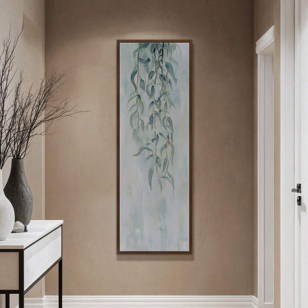 This vertical canvas wall art features a loose, impressionistic painting of pale blue and green willow branches against a textured light blue background, framed in dark wood and displayed on.