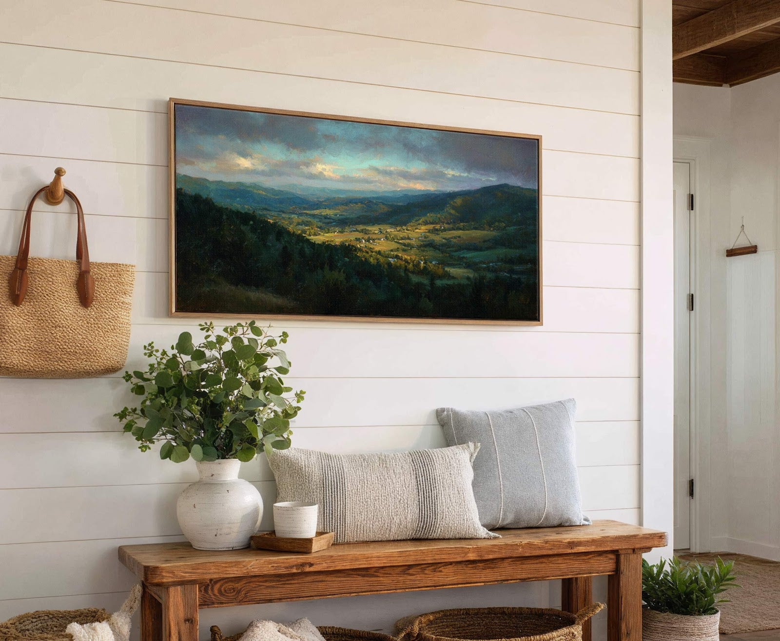 This horizontal canvas wall art features a painterly landscape with a village nestled in a valley under a dramatic, cloudy sky, set against a white paneled wall above a wooden.