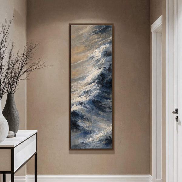 A vertical canvas print features an abstract painting of crashing waves with a palette of deep blues, white, and gold, set against a textured tan wall above a console table.
