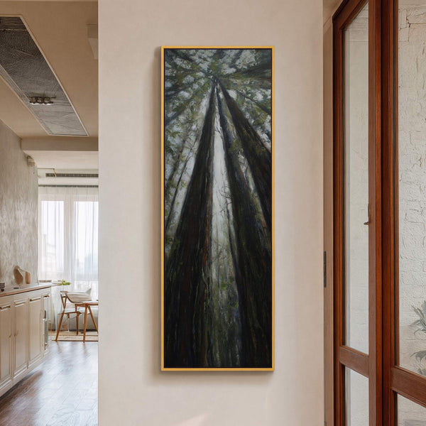 This vertical canvas wall art features a dark, atmospheric painting of redwood trees stretching upwards, framed in gold. It is displayed on a light-colored wall in a room with a.