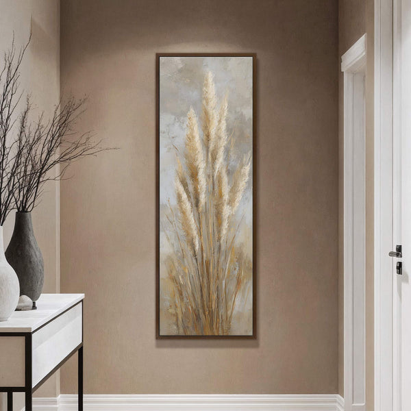 This vertical canvas print features an impressionistic painting of pampas grass in a warm, neutral palette, framed in a dark wood floater frame, hanging on a taupe wall above a.