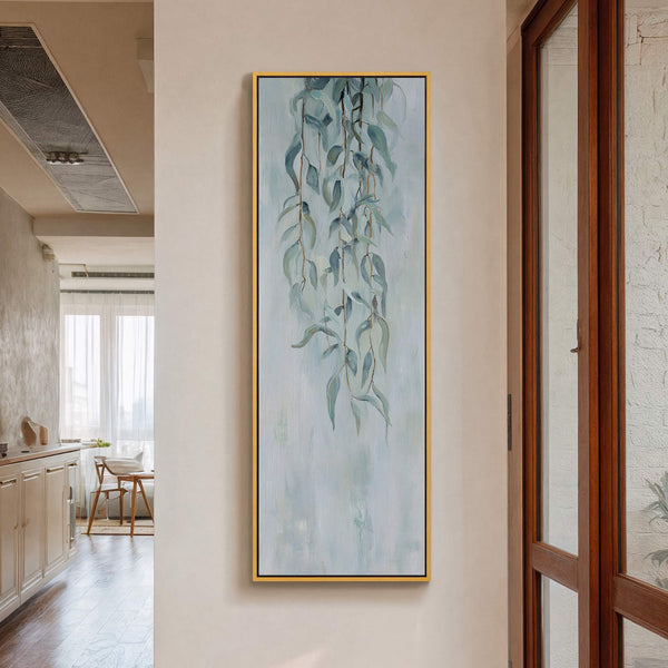 This vertical canvas wall art features an abstract painting of light blue eucalyptus leaves against a textured pale blue background, framed in gold, hanging on a beige wall in-door wall.