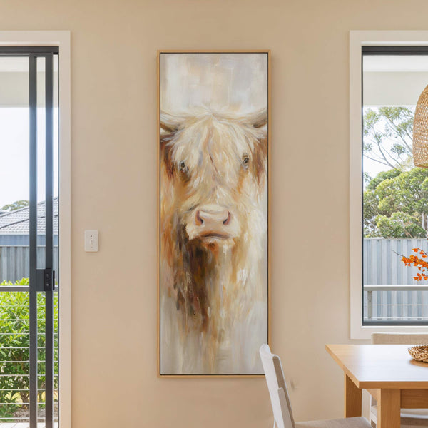This vertical canvas print features a close-up, impressionistic painting of a shaggy Highland cow's face in earthy tones and cream, framed in natural wood, hanging on a beige wall beside.