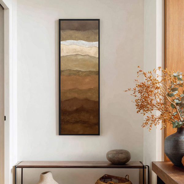 This vertical, abstract canvas wall art features a warm earth-toned palette and is framed in black. It hangs on a textured cream wall above a wooden console table with a.