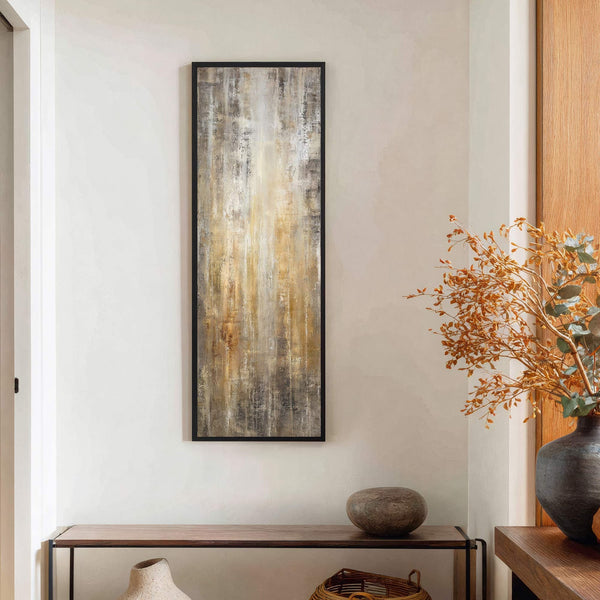 This vertical abstract art print features a palette of gold, brown, and gray tones with a textured, streaky style. It is framed in black and hangs on a light-colored wall.