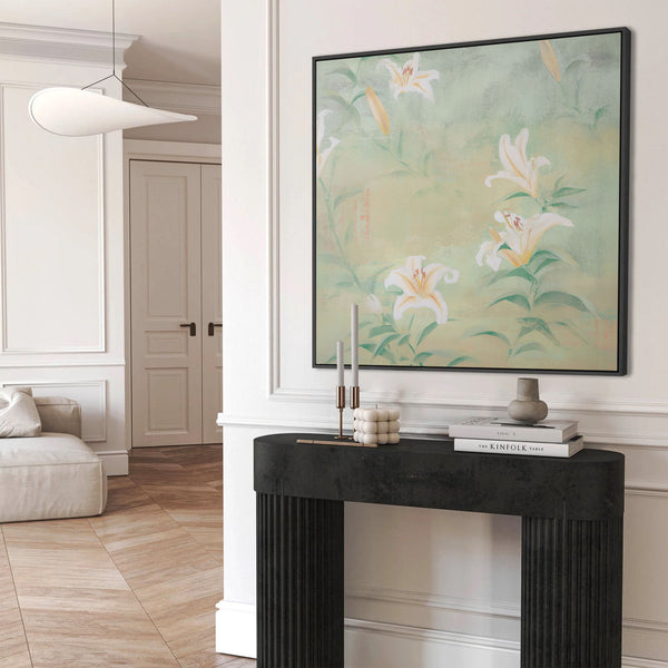 Large black-framed tranquil white lily botanical art print featuring elegant white and yellow lilies on green stems against a soft, muted green and cream textured background, displayed above a black console table in a minimalist white entryway.
