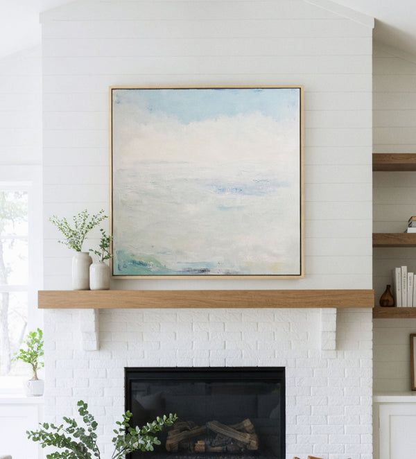 Large natural framed abstract landscape art print featuring soft, layered shades of blue, white, and cream resembling a misty sky and distant horizon, displayed above a white brick fireplace with a natural wood mantel in a bright living room.