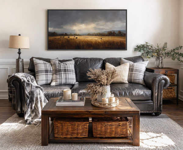 This horizontal canvas wall art displays a dark, moody landscape painting of hay bales in a golden field under dramatic clouds, framed in black, and hung above a dark leather.