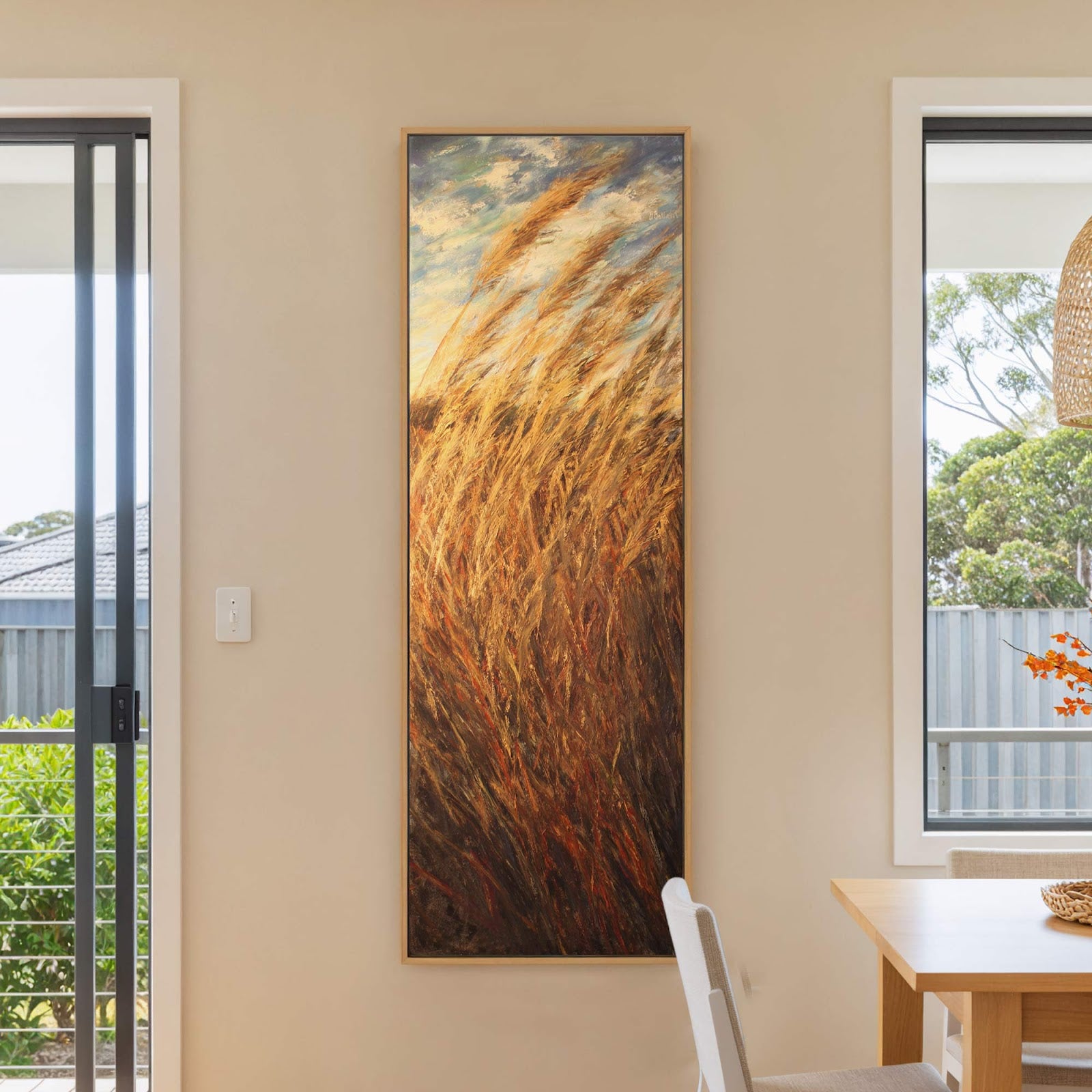 This vertical canvas wall art depicts dry grasses in warm, earthy tones with expressive brushstrokes, displayed in a natural wood floater frame on a cream-colored wall next to a large.